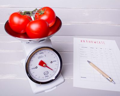 Auf einer Küchenwaage werden Tomaten abgewogen. Daneben liegt ein Zettel mit einer Tabelle, wo Nährwerte von Lebensmitteln eingetragen werden.