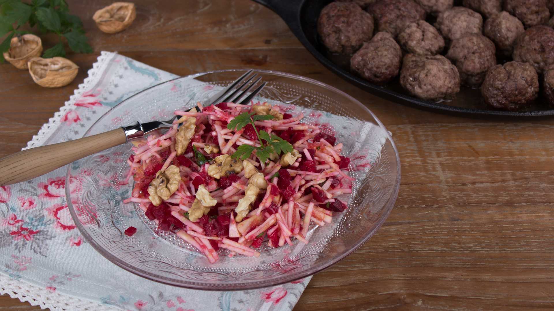 Auf einem Glasteller ist ein Sellerie-Rote-Bete Salat angerichtet, im Hintergrund ist eine Pfanne mit Hackbällchen